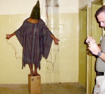 A soldier checks a digital camera while an Abu Ghraib prisoner stands connected to a battery.